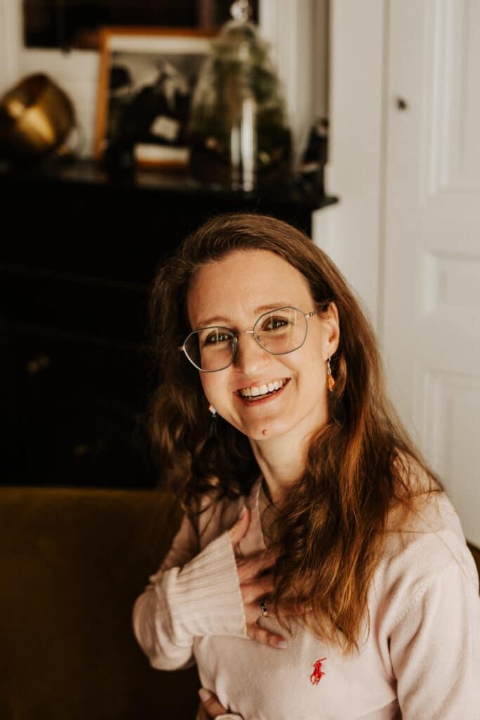 consultante en lactation souriante, assise avec une main sur le cœur et l'autre sur le ventre dans une ambiance douce et chaleureuse -spécialisée dans l'accompagnement des mères après un deuil périnatale, une PMA ou un accouchement difficile.